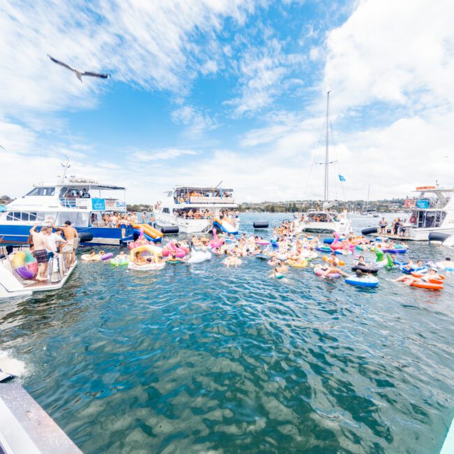 A lively scene of a floating beach party with people on various colorful inflatable rafts in the water. Boats are anchored nearby and seagulls are flying overhead under a partly cloudy sky, accompanied by the rhythmic sounds of music creating an energetic atmosphere.