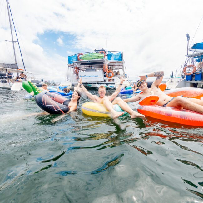 Four people are relaxing on inflatable floaties in the water near anchored boats, smiling and enjoying themselves. In the background, several others are on boats under a partly cloudy sky.