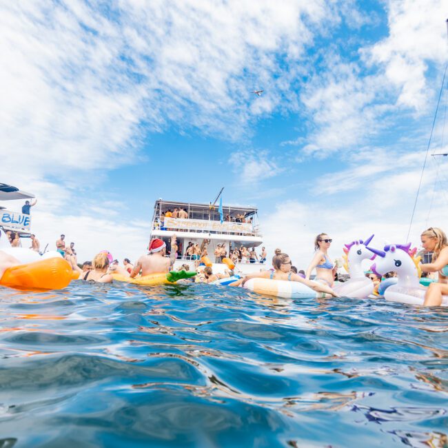 People are floating on inflatable pool toys, including unicorns and flamingos, in the water near boats. The sky is partly cloudy, and a mix of sailboats and motorboats are anchored nearby. Everyone appears to be enjoying a lively, sunny day on the tranquil sea.