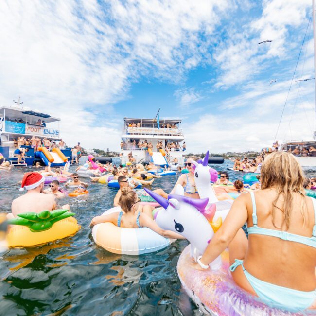 The image shows a vibrant scene of people enjoying a sunny day on the water with colorful inflatable floats. Boats are in the background, and many are using slides from the boats. The festive atmosphere features unique floats, including unicorn and flamingo designs, amidst surfers riding gentle waves.