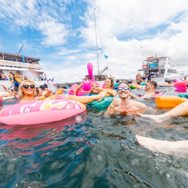 A lively group of people enjoy a sunny day in the water, floating on colorful inflatable pool toys, including flamingos and donuts. Smiling individuals are having fun near a charming beach house in the background under a blue sky with scattered clouds.