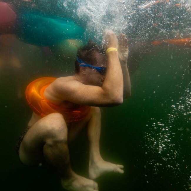 A person wearing blue goggles and an orange swim ring is underwater, crouched with arms raised over the head. Bubbles surround them as other swimmers and floating objects are visible in the background. The scene captures the essence of a fun Yacht Social Club outing.