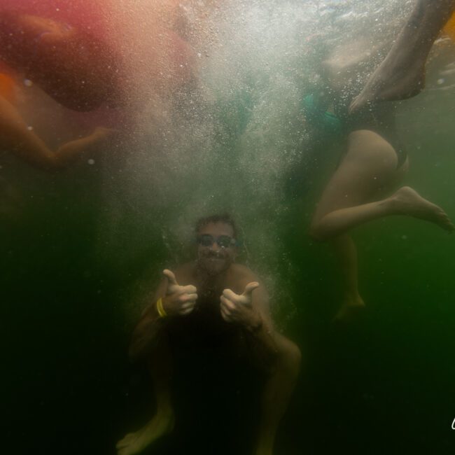 An underwater photo shows a person giving a thumbs-up and smiling amidst bubbles and other swimmers' legs. The water has a greenish hue, and the scene feels playful and fun. The "Yacht Social Club" logo is visible in the bottom right corner, adding an exclusive touch to the joyful moment.