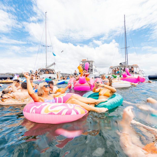 A lively scene of people enjoying a summer day on the water, floating on colorful inflatable rings and rafts. Boats from The Yacht Social Club are anchored in the background. The sky is partly cloudy, and everyone appears to be having fun and relaxing.