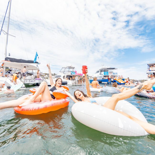 Two young women float on inflatable rings, one orange and one white, in a calm body of water. Both are smiling and posing with arms raised, one holding a red cup. Boats are anchored in the background under a partly cloudy sky. Text reads "Yacht Social Club" in the bottom corner, emphasizing fun on the water.