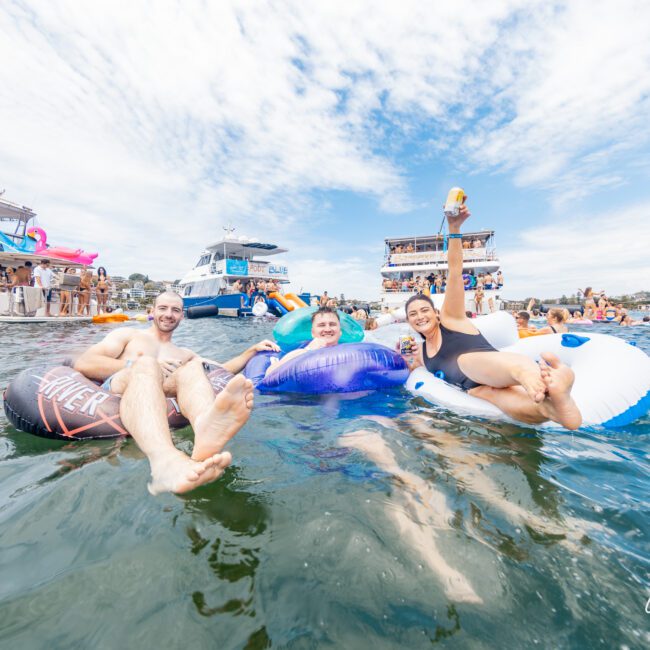 Three people are floating on inflatable rafts in the water, smiling and enjoying a sunny day. They are surrounded by other people on kayaks and boats. One person is raising a drink. Boats are anchored in the background with partygoers visible. The atmosphere is festive and vibrant.