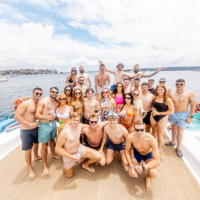 A large group of men and women in colorful summer attire and swimsuits pose together on a boat deck with the ocean and blue sky in the background. They appear happy, with some raising their hands in celebration. The picturesque shoreline with buildings is visible in the distance.