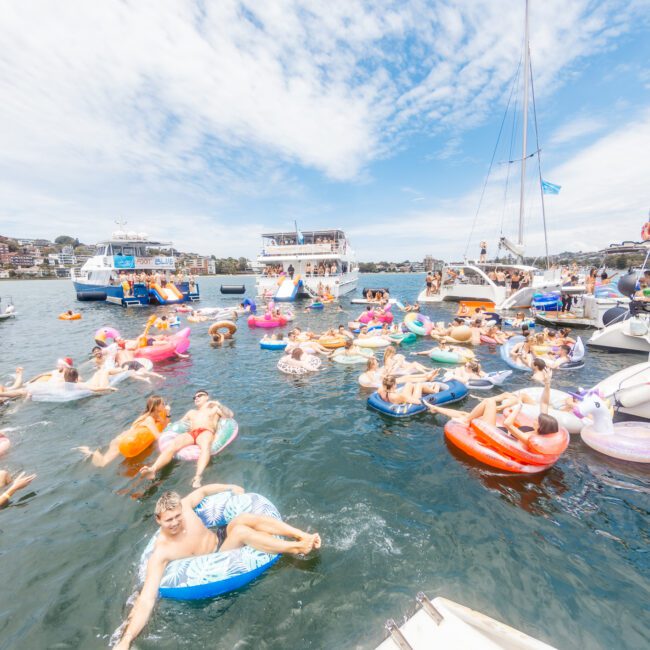 A sunny day on the water shows a lively scene with many people enjoying themselves on vibrant inflatable floats. Various boats are anchored nearby, and numerous individuals are seen swimming and relaxing against a backdrop of partly cloudy skies and charming coastal buildings.