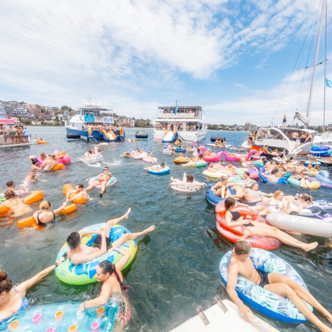 A vibrant gathering of people floating on colorful inflatable tubes and rafts in a sunny harbor creates a festive atmosphere. Boats are docked in the background, under a partly cloudy sky. Everyone appears relaxed, soaking up the sun and enjoying the lively waterfront scene.