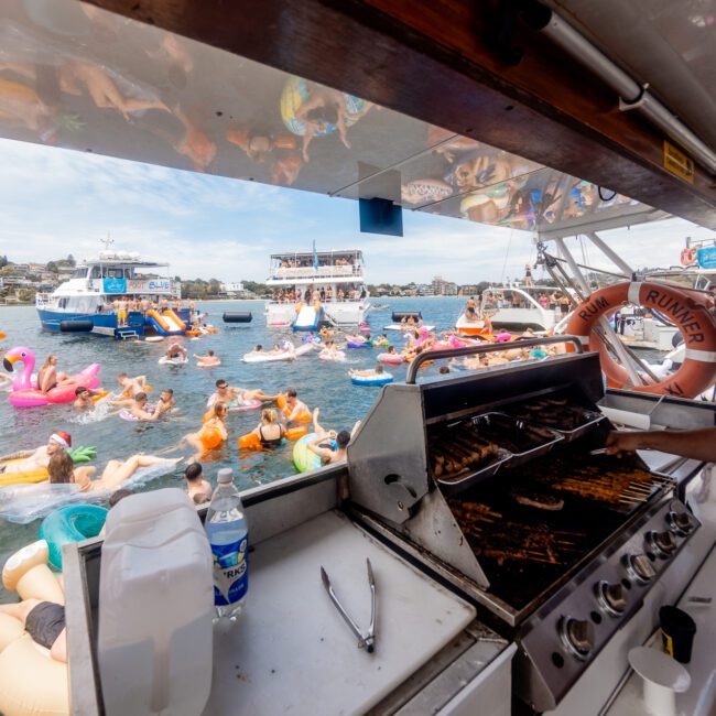 A person grills food on a boat surrounded by water with numerous colorful, inflatable floats and people swimming. In the background, several boats are anchored under a clear sky with some clouds. The atmosphere appears lively and festive, perfect for paddleboarding enthusiasts.