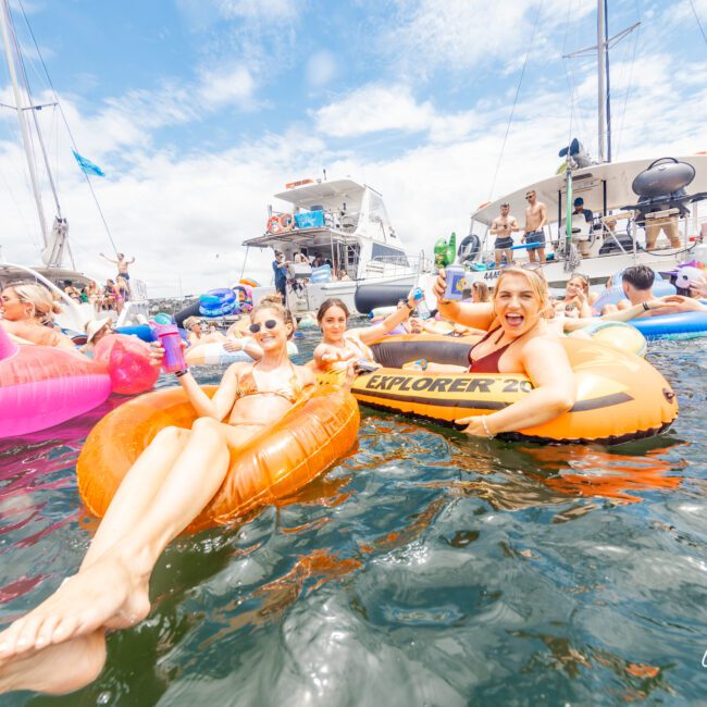 A lively group of people enjoys a sunny day on the water, floating on colorful inflatables near docked yachts. Some hold drinks while others smile at the camera. Bright inflatables including flamingos and donuts add to the fun. The water is calm and the sky is partly cloudy, creating an inviting atmosphere.