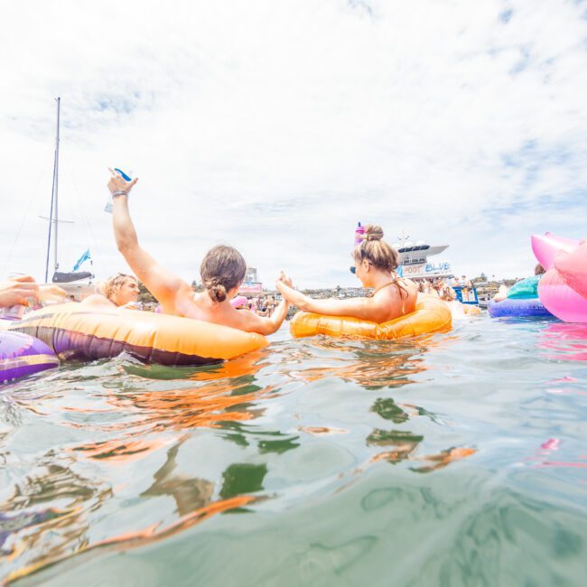 Two people enjoying a sunny day on a body of water, relaxing on inflatable tubes under the clear sky. They are reaching out to each other with boats visible in the background. The person in the orange tube is raising their drink, embodying the carefree vibe of Yacht Social Club.