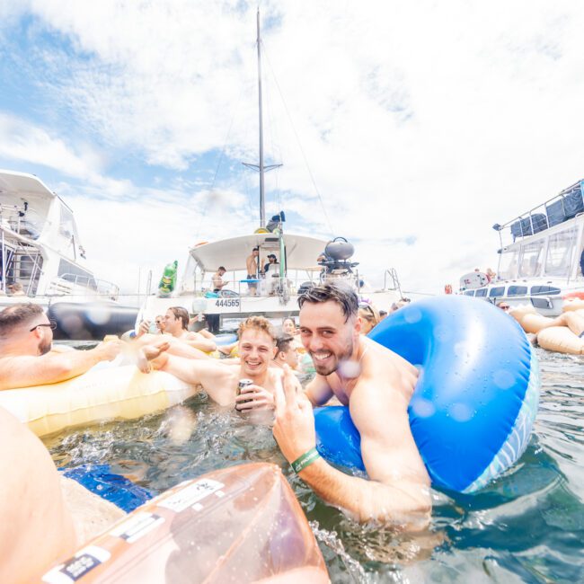 A group of people enjoying a sunny day on the water, floating on inflatable tubes. One man in the foreground floats in a blue tube, smiling and holding a drink. Boats and yachts are visible in the background under a partly cloudy sky, creating a vibrant summer scene filled with joy and relaxation.