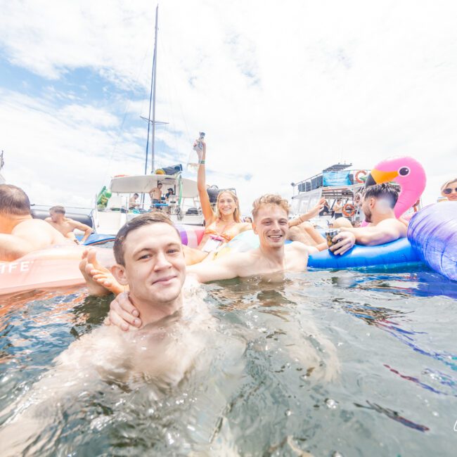 A group of friends enjoying a sunny day in the water near several yachts. Two men in the foreground smile at the camera while others relax on floaties, including a pink flamingo. One woman holds up a drink under the partly cloudy sky. The picturesque scene is perfect for capturing memories.