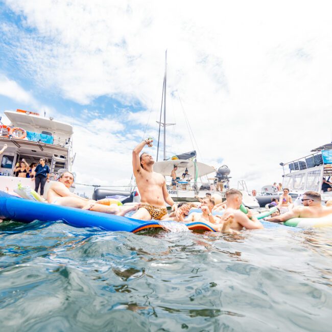 A lively group of people enjoying a boat party on a sunny day. Many are on large inflatables in the water, socializing and holding refreshing drinks. Several yachts are anchored in the background, their reflections shimmering. The atmosphere is festive and relaxed, with music enhancing the joyous mood.