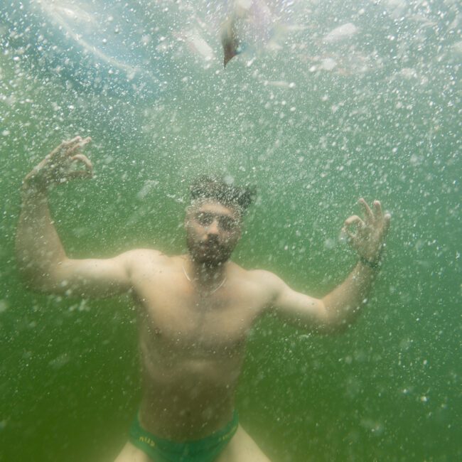 An underwater photo captures a man wearing green swim trunks, submerged with bubbles creating an enchanting effect. He's posing confidently with his hands in front, fingers forming an "OK" sign. The prestigious Yacht Social Club logo is visible in the backdrop.