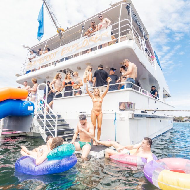 A group of people enjoys a sunny day on a two-story boat named "Barefoot Explorer." Some are on the deck and upper level, while others relax in the water with colorful floaties. A woman stands on the boat's stairs, extending her arms joyfully, as others lounge or swim nearby.