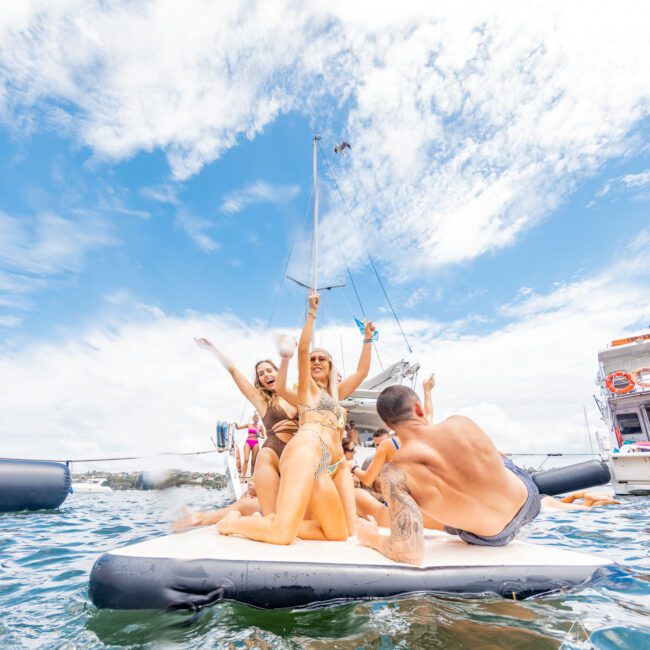 A group of people in colorful swimwear enjoying themselves on a floating inflatable platform in the water. They are smiling, posing, and making celebratory gestures with a boat in the background under a partly cloudy sky.