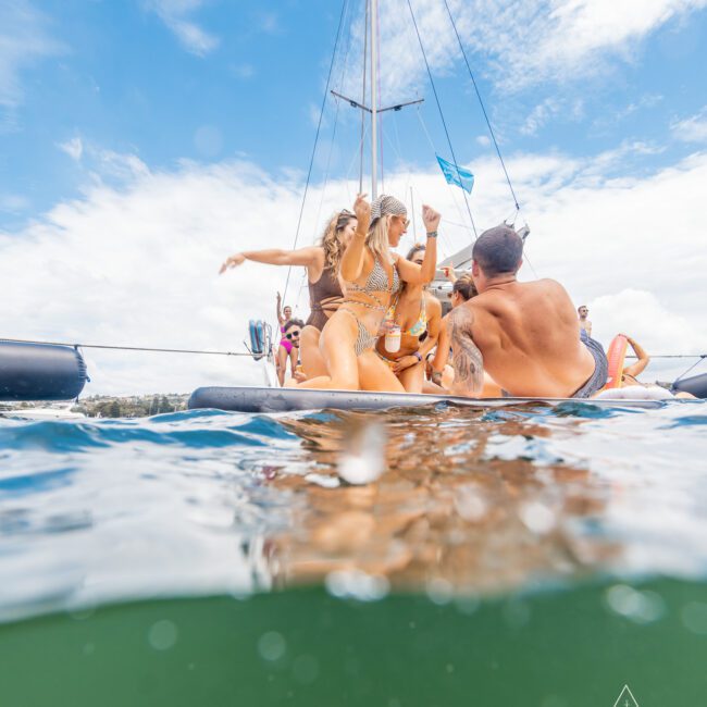 A group of people in swimsuits enjoying a yacht party, some standing and some sitting, with a mast and flag visible in the background. The photo, capturing the fun just above the water's surface, shows a partly cloudy sky. The logo in the bottom right reads "The Yacht Social Club".