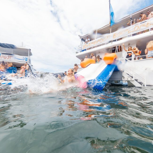 A vibrant scene of people enjoying themselves on a large boat with water slides leading into the ocean. Some individuals are sliding down, creating splashes, while others are standing or sitting on the boat’s multiple levels, socializing and watching the lively activity unfold.