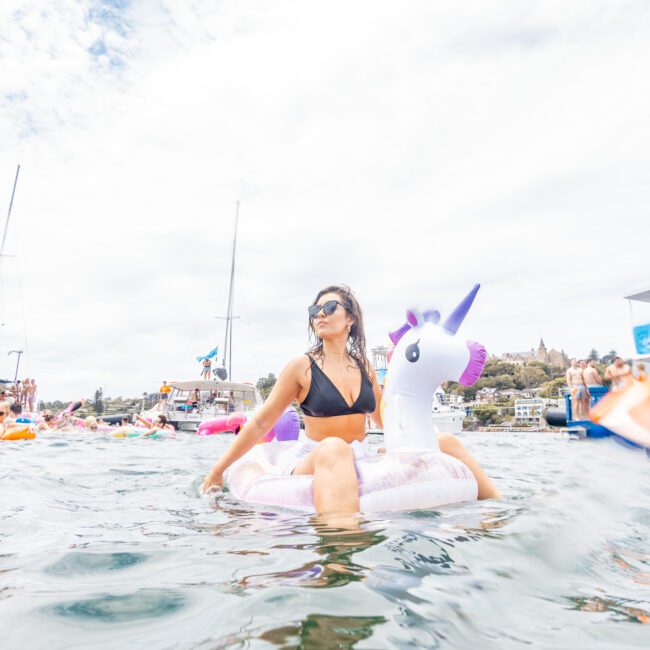 A person in a black bikini rides a white and purple unicorn float in a large body of water, surrounded by others on various inflatables and boats. The sky is partly cloudy, with greenery and buildings visible in the background, giving the scene a fun and whimsical atmosphere.