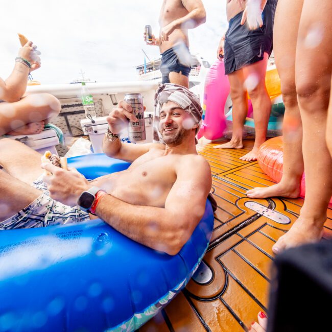 A group of people is enjoying a vibrant pool party on a boat. One person lounges in an inflatable blue ring, holding a drink and smiling, while others stand nearby in swimsuits, some holding drinks. The atmosphere is lively and fun, with everyone relishing the sunshine and cool water.