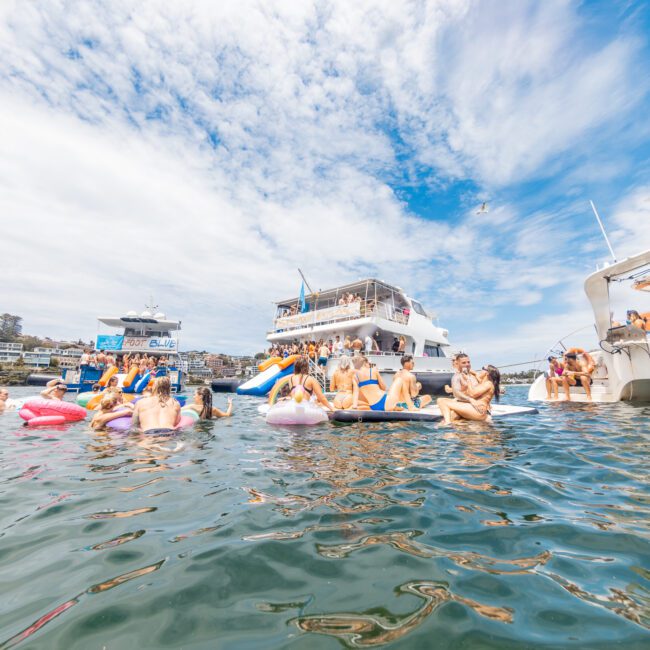 A crowded scene with people enjoying a sunny day on the water, swimming and lounging on inflatables between boats. In the background, buildings can be seen on the shore. The foreground showcases a lively atmosphere of swimmers and people on yachts from Sydney Harbour Boat Hire The Yacht Social Club.