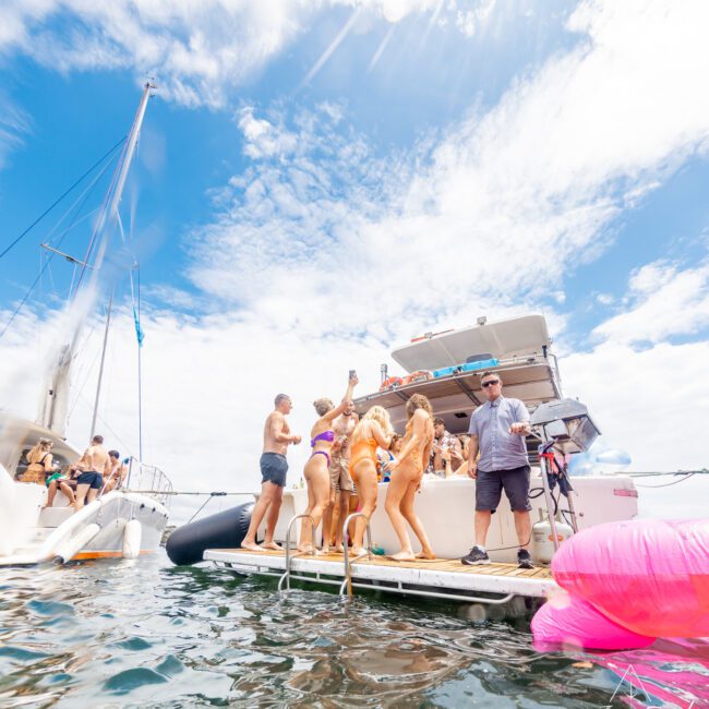 A vibrant scene of several people enjoying a sunny day on a boat. Some are dancing and interacting near the boat's edge, while one person stands on a lower deck with a lifebuoy and inflatable swan nearby. The water is calm, the sky is partly cloudy, and music adds to the festive atmosphere.