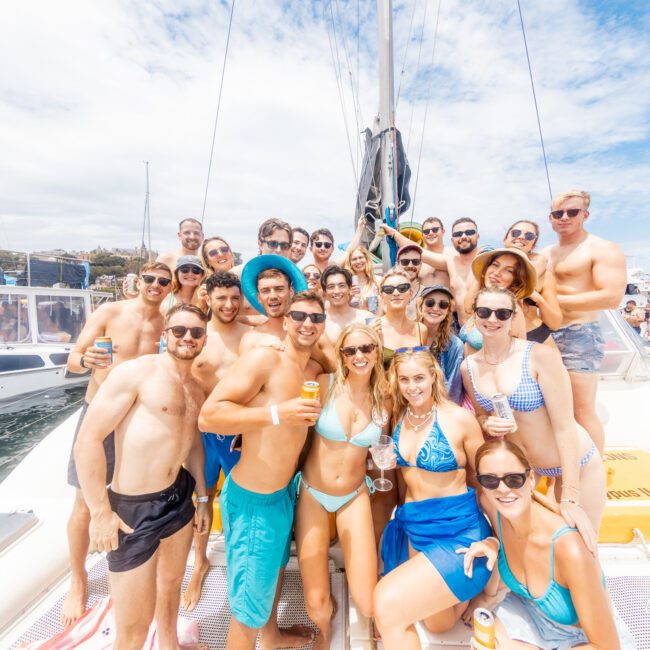 A large group of people gather on a boat, smiling and posing for a photo. Many are wearing swimsuits and sunglasses, with some holding drinks. The sky is partly cloudy, and sailboats can be seen in the background, indicating a lively social atmosphere on the water.