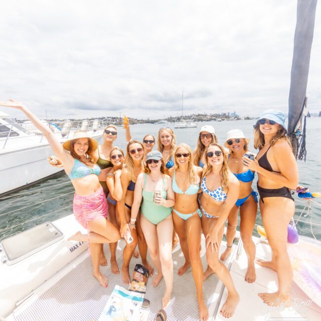 A group of 11 women in stylish swimsuits are gathered on a boat, smiling and posing for a photo. They are enjoying a sunny day on the water, with a city skyline and other boats visible in the background. The mood is cheerful and celebratory, capturing the essence of summer fun.