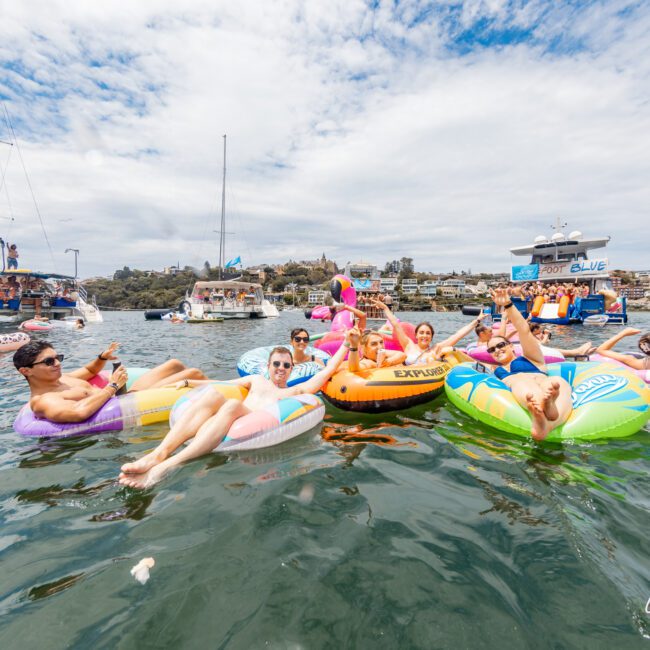 People relax on colorful inflatable tubes in the water during a sunny day, with yachts anchored nearby. The sky is partly cloudy, and in the background, a shoreline with trees and buildings can be seen. A slight breeze ripples across the water, adding to the idyllic scene. A "Yacht Social Club" watermark is in the bottom right corner.