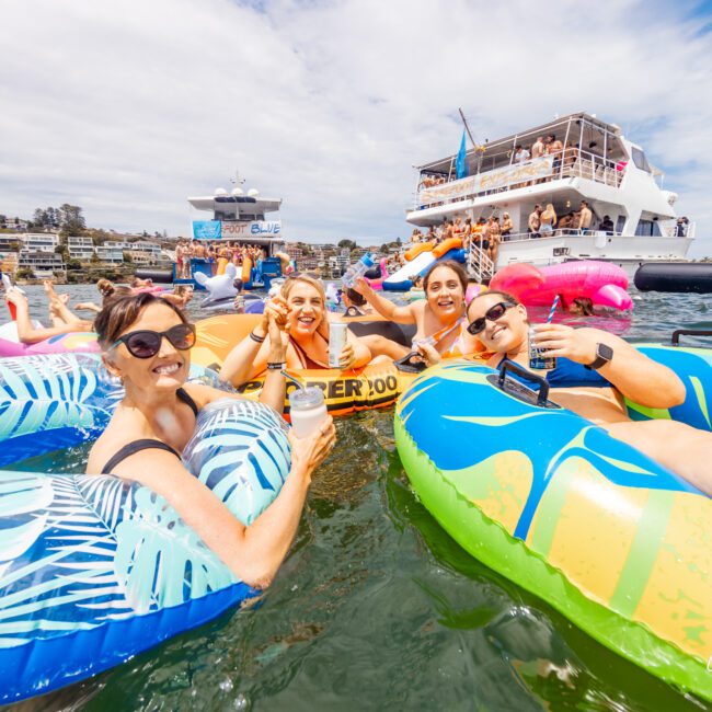 Four people are relaxing in colorful inflatable tubes on a sunny day, each holding drinks and smiling toward the camera. In the background, The Yacht Social Club Event Boat Charters hosts a lively boat party with many attendees, set against a clear blue sky and calm water.