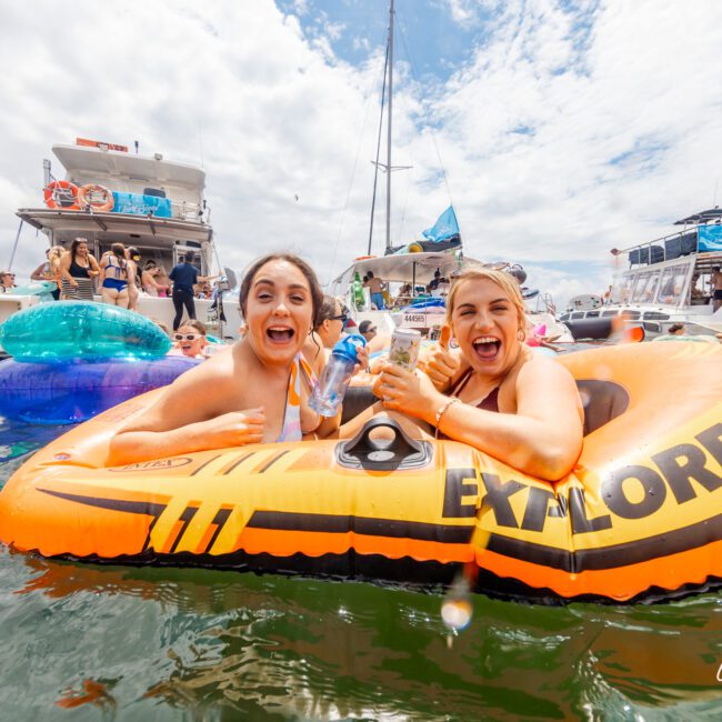 Two women in vibrant swimsuits are joyfully floating on an orange inflatable raft labeled "Explorer 200" in a sunny, bustling marina. Surrounded by others on various floats, they enjoy the lively atmosphere, with several yachts anchored in the background under a partly cloudy sky.