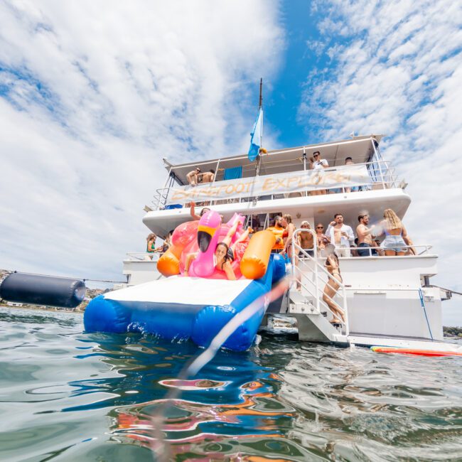 A vibrant boat party scene shows people enjoying themselves on the deck of a large yacht under a partly cloudy sky. Adorned with colorful decorations and floats, the yacht features two people gleefully sliding off an inflatable slide into the inviting water.