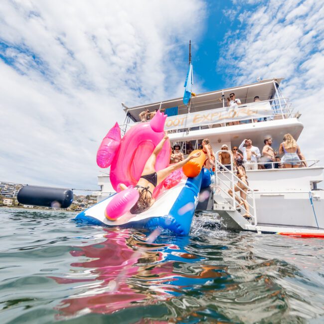 A fun scene of people on a yacht with several individuals in swimsuits enjoying a sunny day. In the foreground, someone is climbing an inflatable slide shaped like a pink flamingo, leading into the calm water. The atmosphere is perfect for relaxation and enjoyment under the partly cloudy sky.