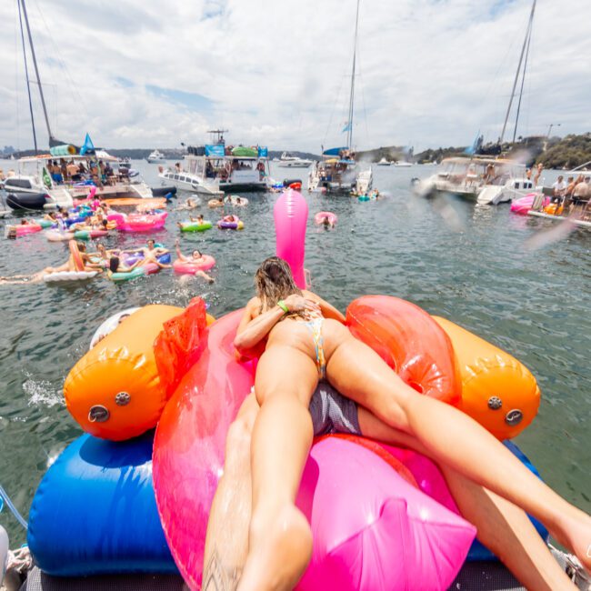 A group of people enjoying a sunny day on a lake, with some relaxing on colorful inflatable floats. The foreground features a person lying on a large flamingo float. Several boats are visible in the background, and the atmosphere is vibrant and festive under the clear blue sky.