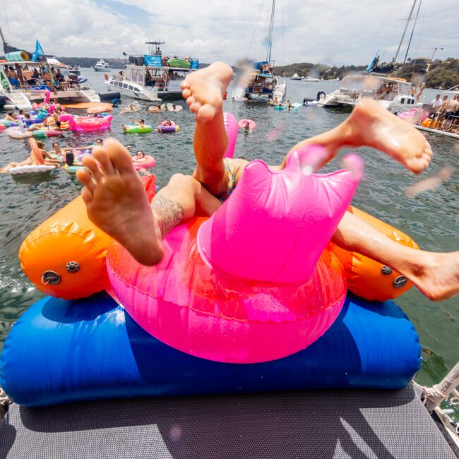 Two people are playfully tumbling on a giant, vibrant inflatable pool toy during a lively boat party on a sunny day. Surrounding them are various boats and colorful inflatables with people enjoying the water. The sky is partially cloudy, adding to the festive atmosphere.