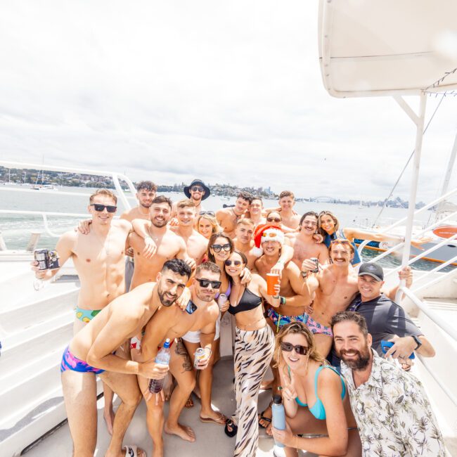 A lively group of people, mostly in swimwear, are gathered on a boat deck, smiling and posing for the camera. The background shows a body of water with a distant shoreline and cloudy sky. The atmosphere is festive, suggesting a fun outing or party hosted by The Yacht Social Club Sydney Boat Hire.