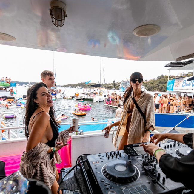 A lively boat party scene with a DJ performing on deck while several people dance and enjoy the music. Various inflatable floats and other boats with numerous partygoers are visible on the water in the background. A banner reading "Yacht Social Club" is in the bottom right corner, adding to the festive atmosphere.