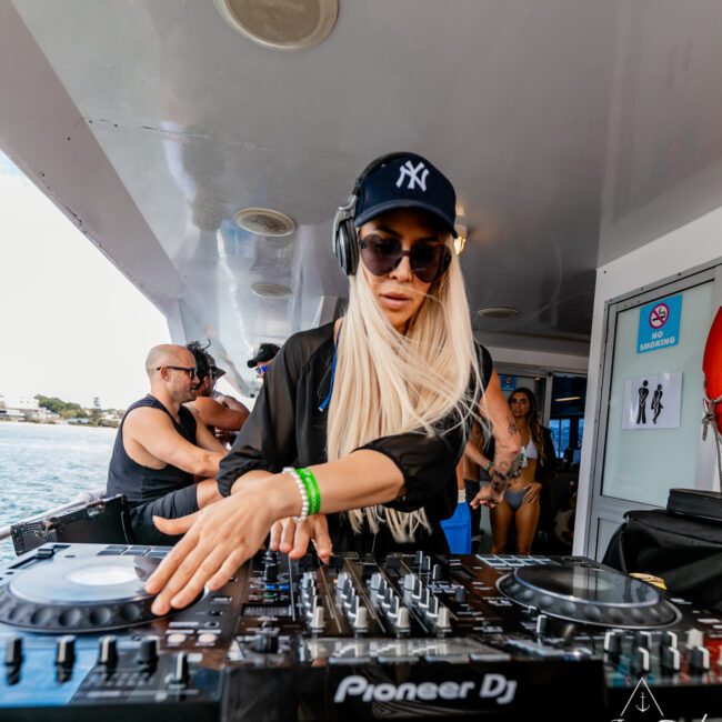 A DJ with long blonde hair, wearing a black cap and sunglasses, is playing music on a Pioneer DJ setup aboard a boat. Other people are seen in the background enjoying the vibrant event under a clear sky, with serene water in view.