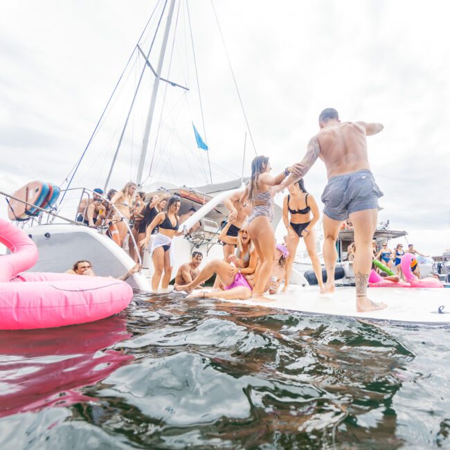 A group of people in swimsuits are gathered on and around a boat in the water. Some are on an inflatable pink flamingo float, while others enjoy relaxing on a white floating platform. One person helps another climb onto the platform, ensuring everyone has fun in the sun.