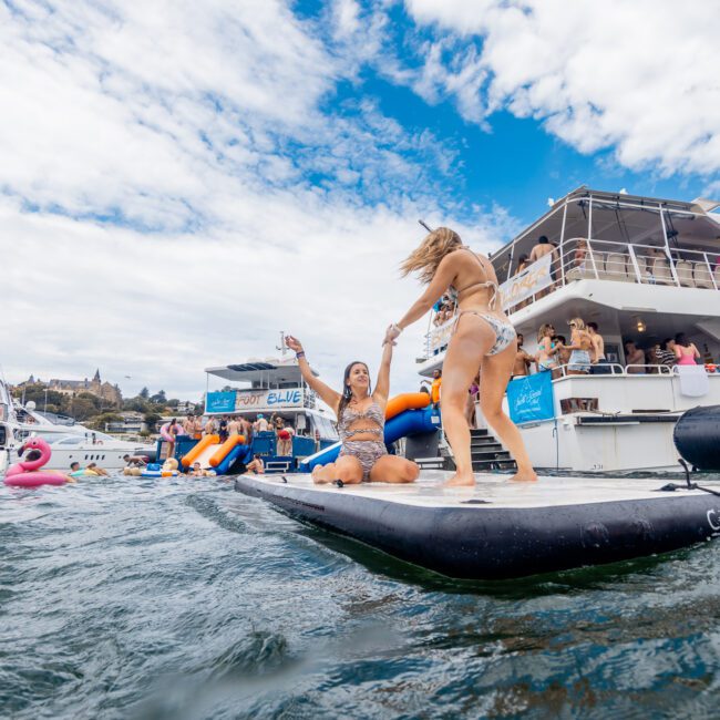 Two women in swimsuits dance on an inflated platform floating on water, surrounded by vibrant boats and inflatables. In the background, several boats and people enjoy the lively atmosphere under a partly cloudy sky.