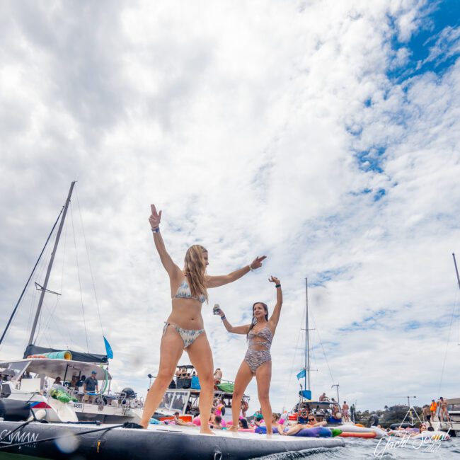 Two women in swimwear stand on an inflatable paddleboard on the water, arms raised and smiling. Boats and other paddleboards populate the background, under a mostly cloudy sky. It appears to be a lively, outdoor water festival filled with joy and excitement.