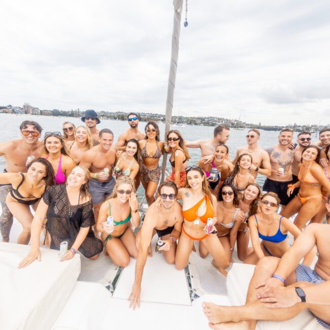 A large group of people in swimsuits and summer attire gather on a boat. They pose for the camera with smiles, making various hand gestures. The background features a crystal-clear body of water and a distant shoreline under a cloudy sky.