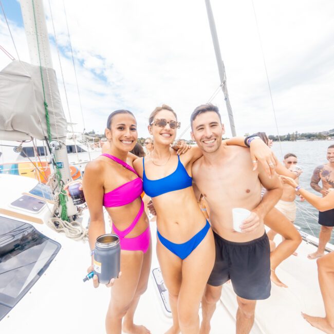 Three people in swimwear smile and pose on the deck of a sailboat. The person on the left wears a vibrant pink bikini, the person in the middle sports a stunning blue bikini, and the person on the right wears stylish black swim trunks. Other people and a picturesque shoreline are visible in the background.