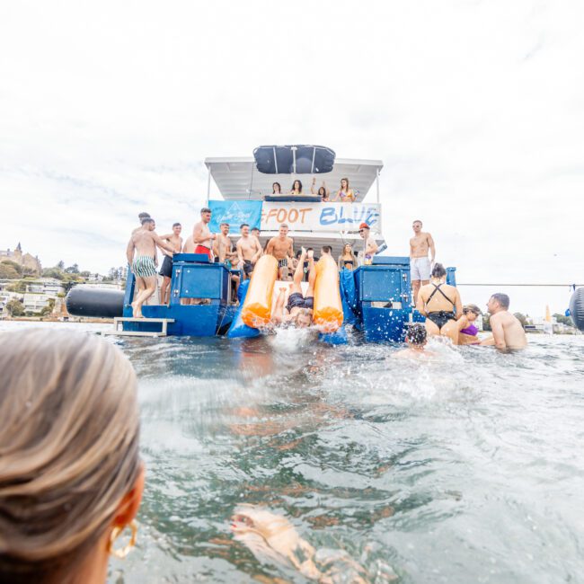 People are enjoying a summer day, swimming around and sliding off a boat with a water slide into the water. The boat has a blue structure with a "Foot Blue" banner. In the background, coastal houses and trees complete the picturesque landscape.