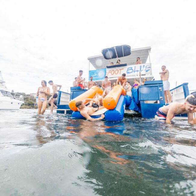 A group of people enjoys a fun day on a boat. Several are sliding down an inflatable slide into the water, while others stand or sit on the deck. In the background, another boat is partially visible. The sky is cloudy, and everyone looks relaxed and happy in this lakeside adventure.