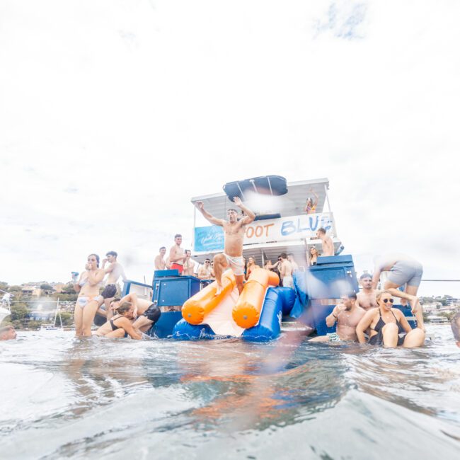 A group of people enjoying a party on a boat in the water, some sitting on the edge while others are partially submerged. Inflatable orange and blue floats add color to the lively and festive atmosphere under a cloudy sky, creating a scene filled with joy and camaraderie.