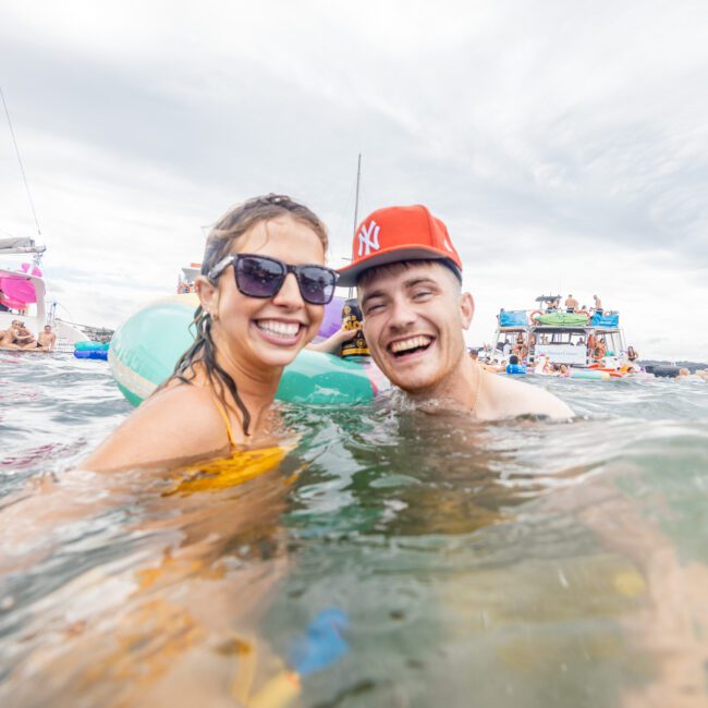 A woman wearing sunglasses and a man in a red cap smile while standing in water. More people and boats are visible in the background, suggesting a lively, fun-filled event on the water. The atmosphere appears joyful and festive.