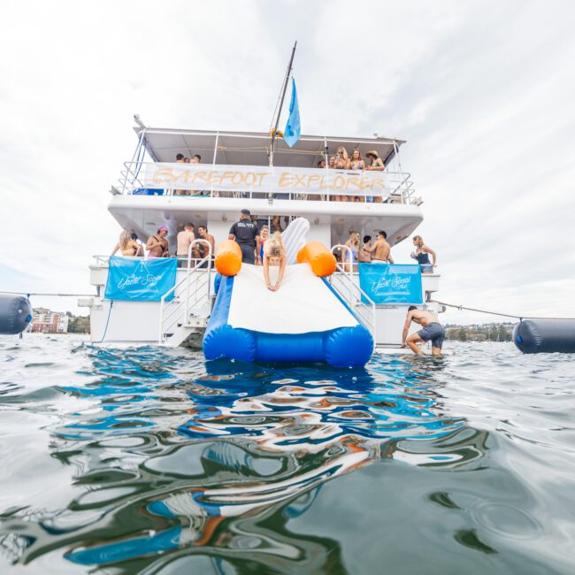 A group of people enjoying a sunny day on a double-deck boat named 'Barefoot Explorer'. The back of the boat features a blue and white inflatable water slide with someone sliding down. The calm, crystal-clear water surrounds the boat, where a few people are happily swimming nearby.